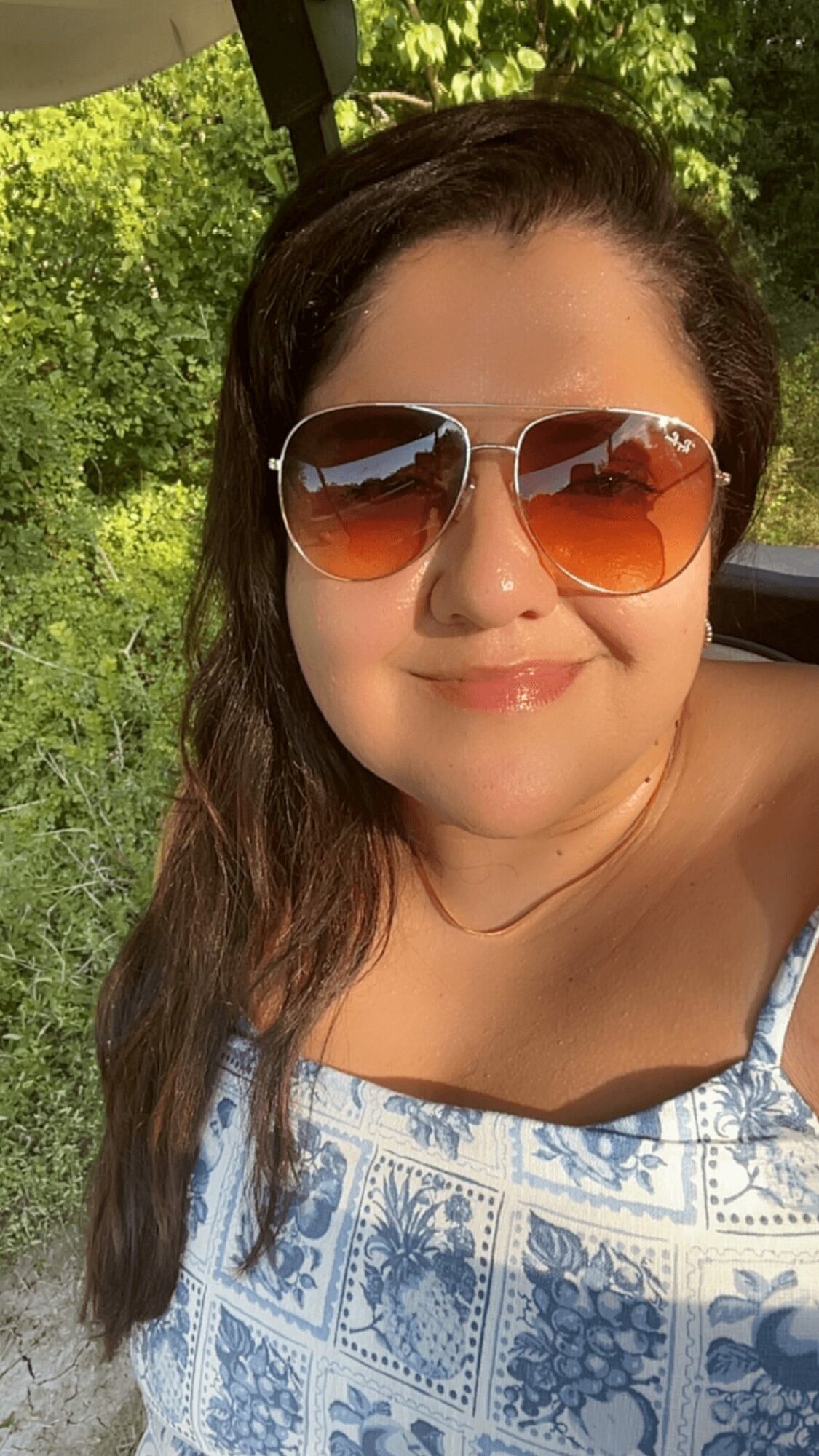 Woman wearing sunglasses and a patterned top outdoors with green trees in background.