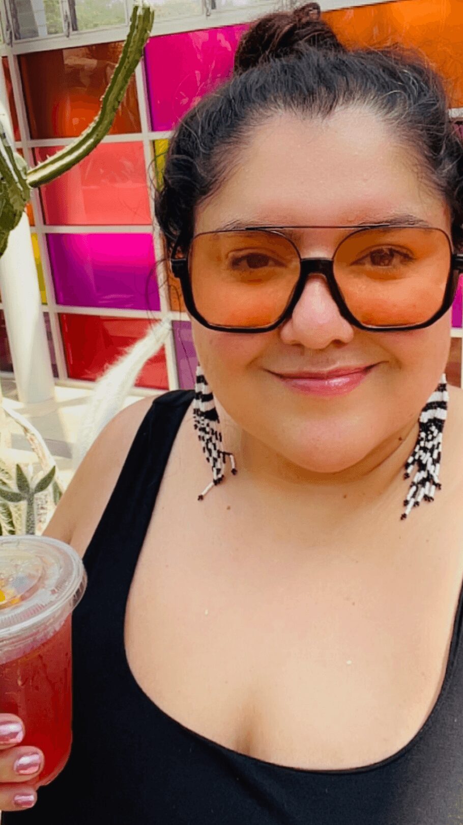Woman with sunglasses holding a red drink, smiling, in front of colorful background and plants.