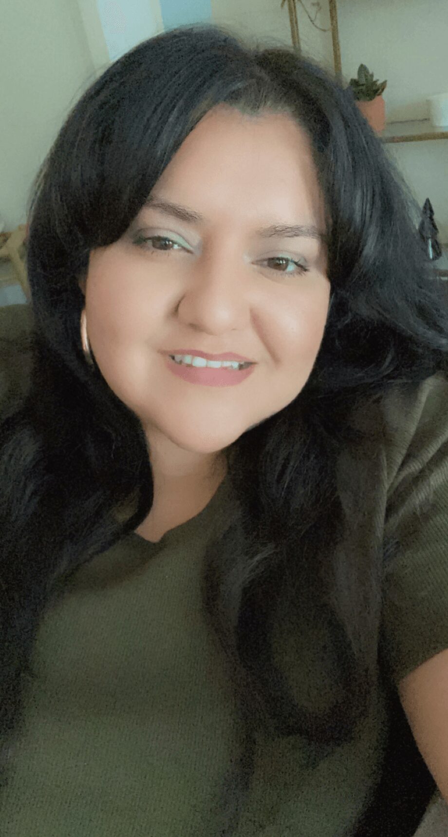 Woman with long dark hair smiling, wearing a green top, in indoor setting with shelves and plants in background.