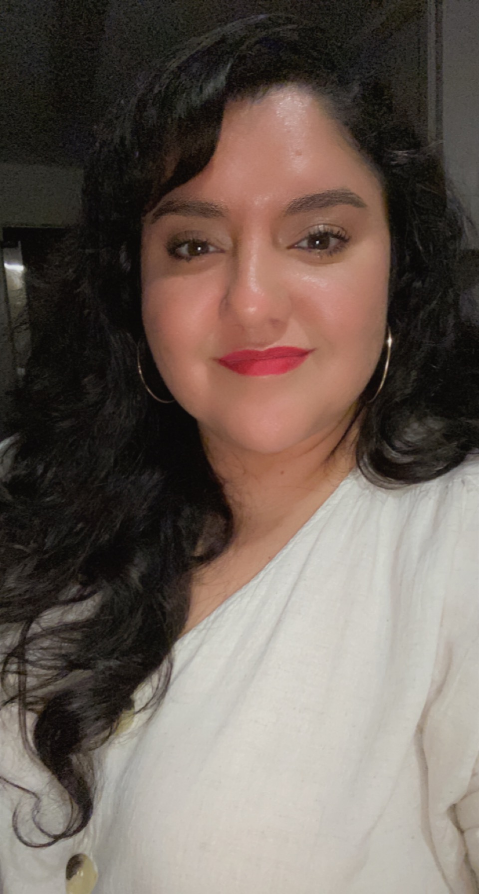 Woman with dark wavy hair, wearing hoop earrings and a white top, smiling at the camera.