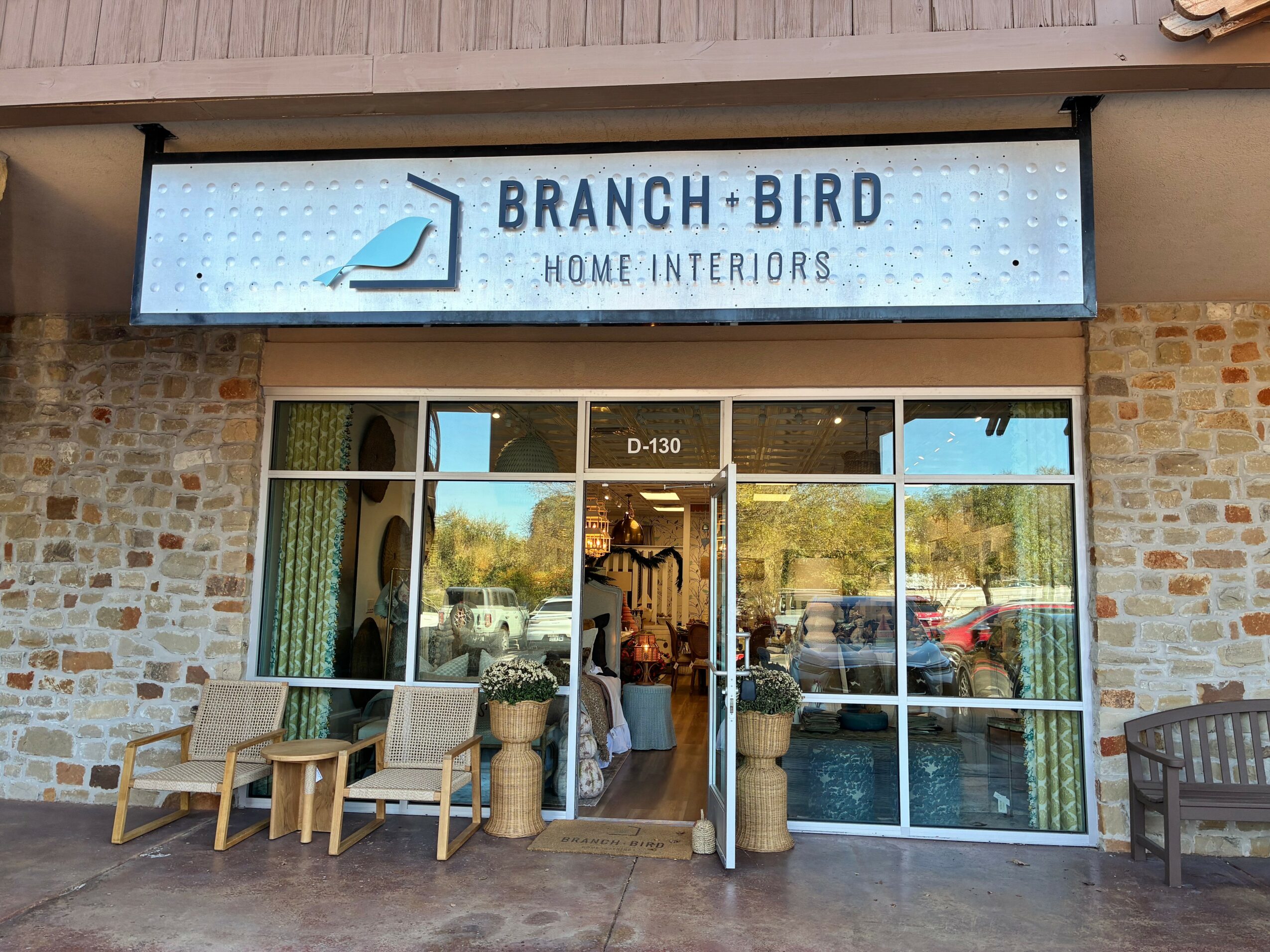Storefront with glass doors and windows, sign reads 'Branch + Bird Home Interiors,' outdoor seating and potted plants.