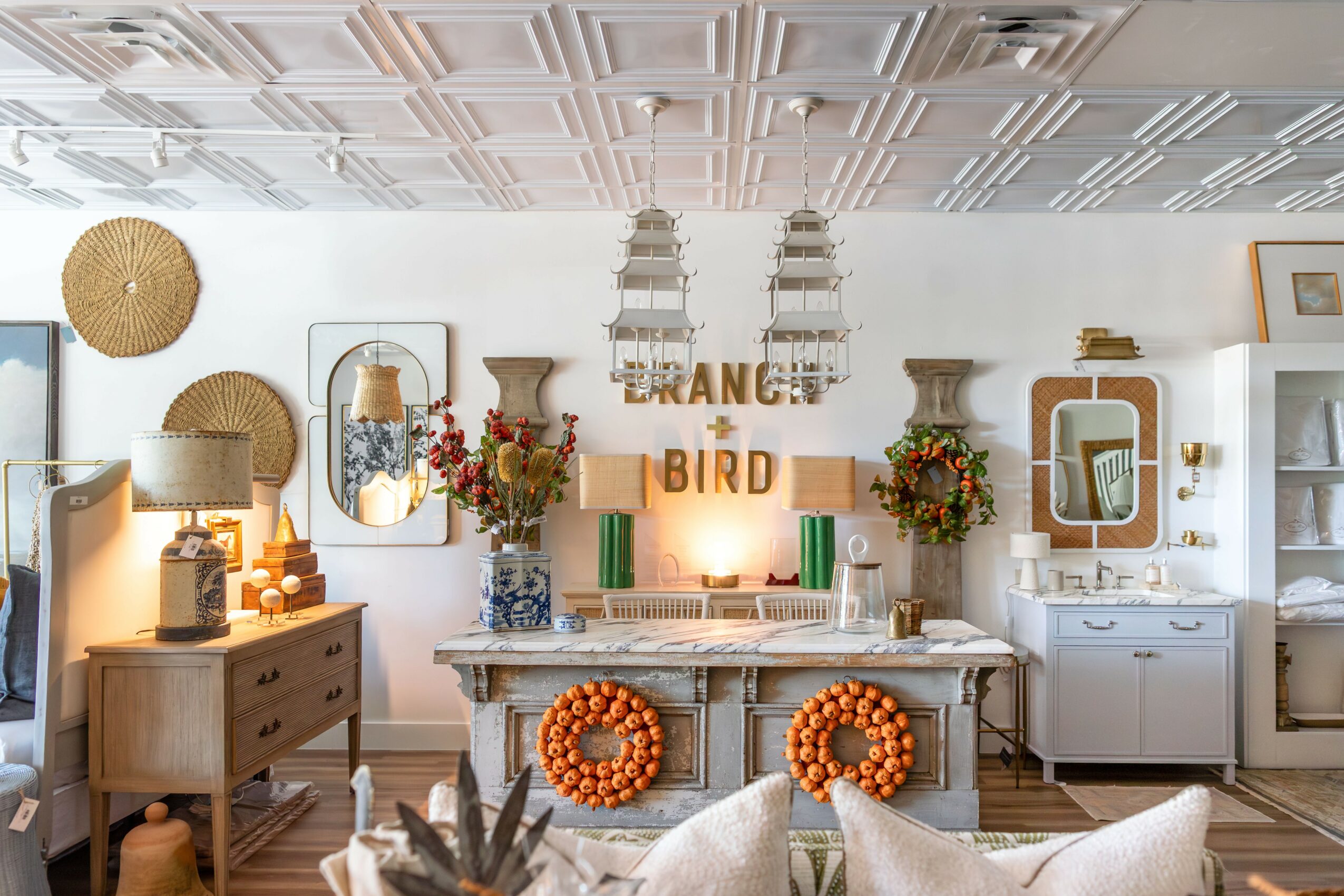 Living room with white walls, decorated with plants, lamps, and wall art, featuring a table with orange wreaths and decorative items.