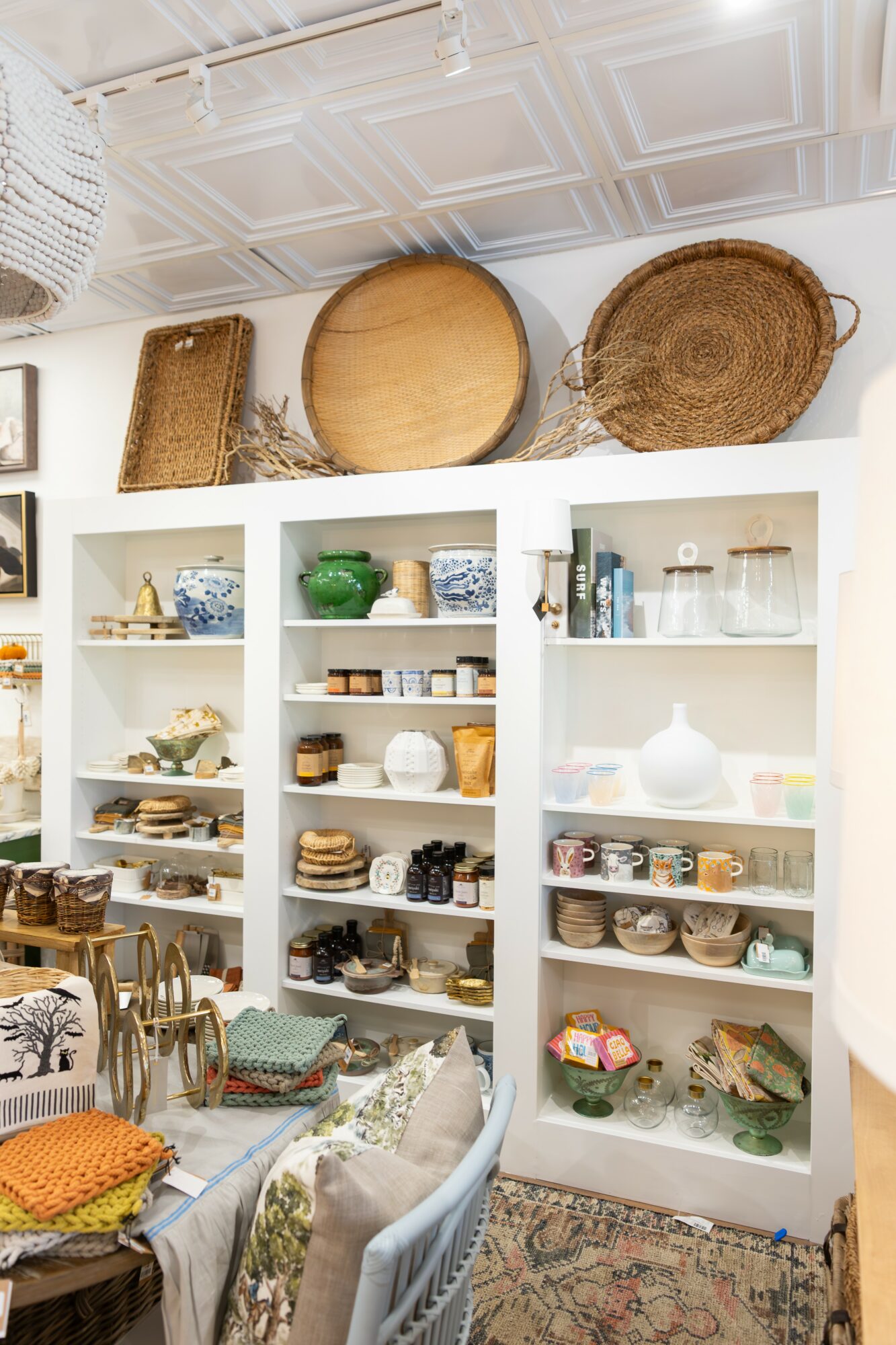 White shelves filled with dishes, bowls, and decorative items in a cozy room with a patterned rug and chairs.