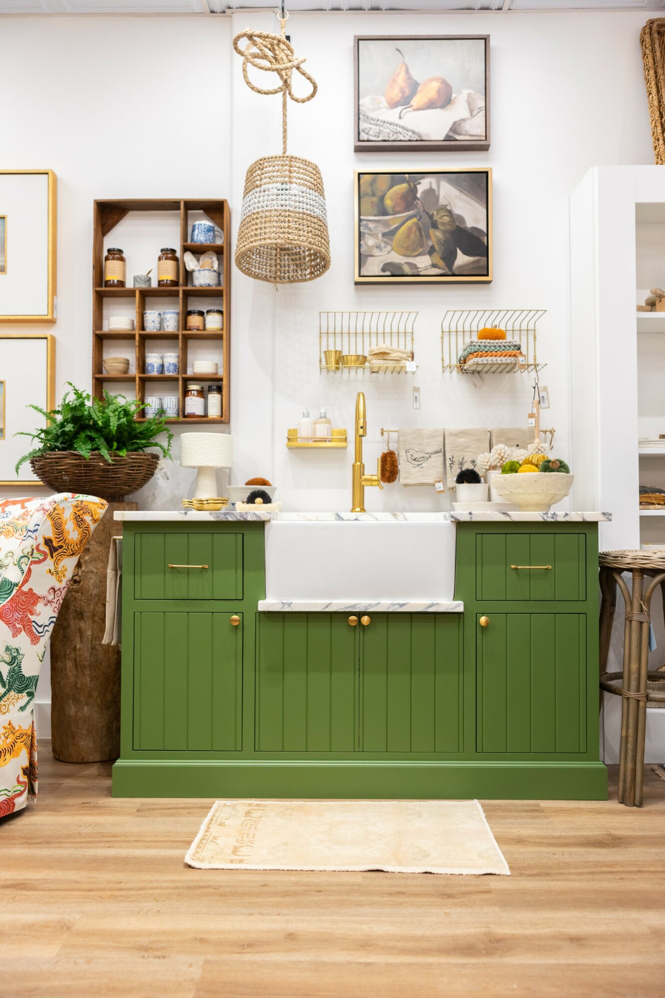 Green kitchen cabinet with white farmhouse sink, wooden countertop, and decorative items. Wooden shelves and framed pictures on wall.