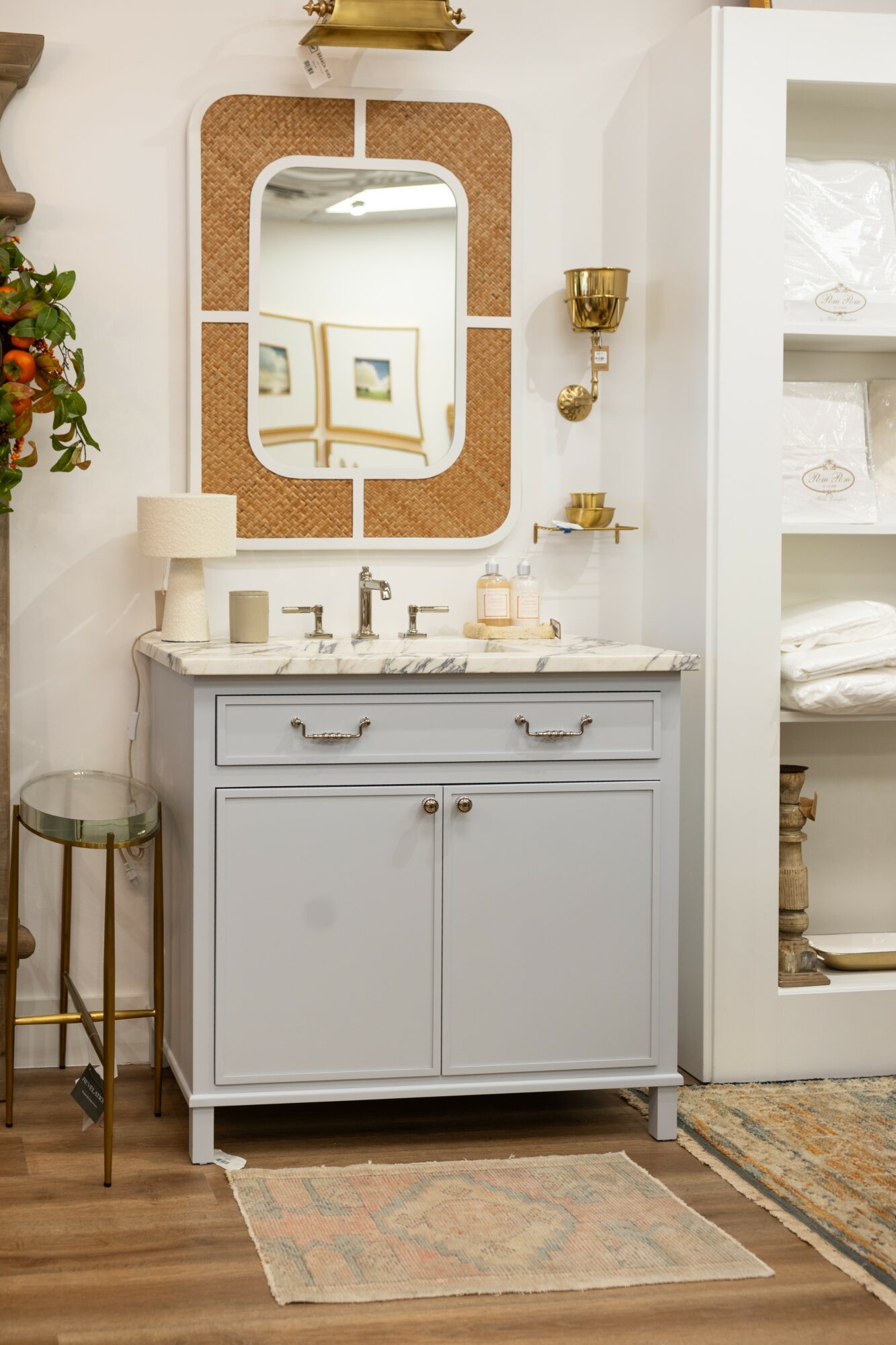 Bathroom vanity with mirror, gold wall sconce, and white cabinet with towels and decor, wooden floor, and rug.