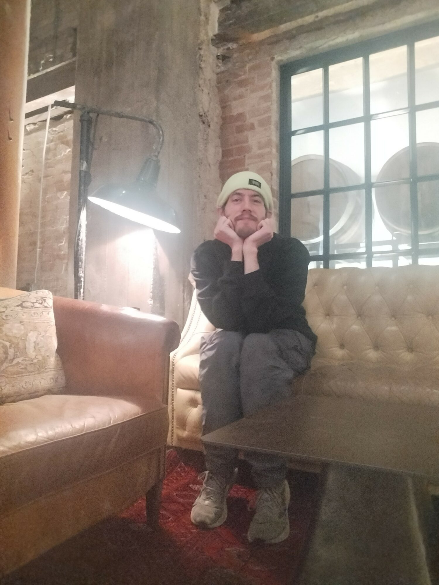 Child sitting on a beige sofa, wearing a white beanie, black jacket, and gray pants, smiling with hands on cheeks.