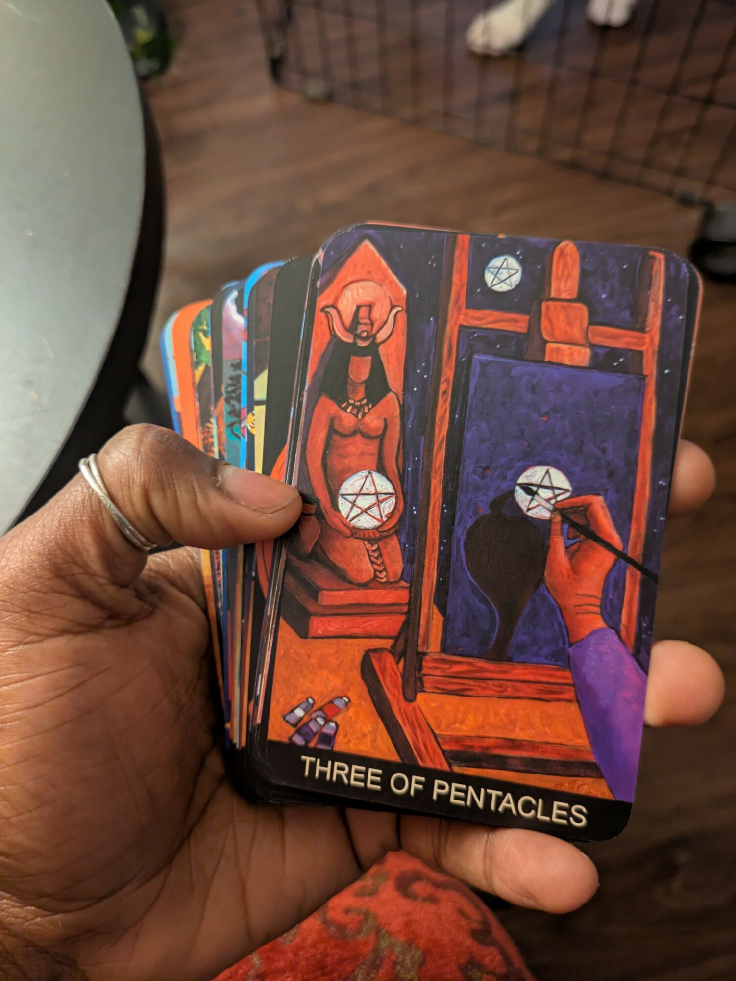 Hand holding tarot cards with colorful illustrations, including a figure and symbols, labeled 'Three of Pentacles'.