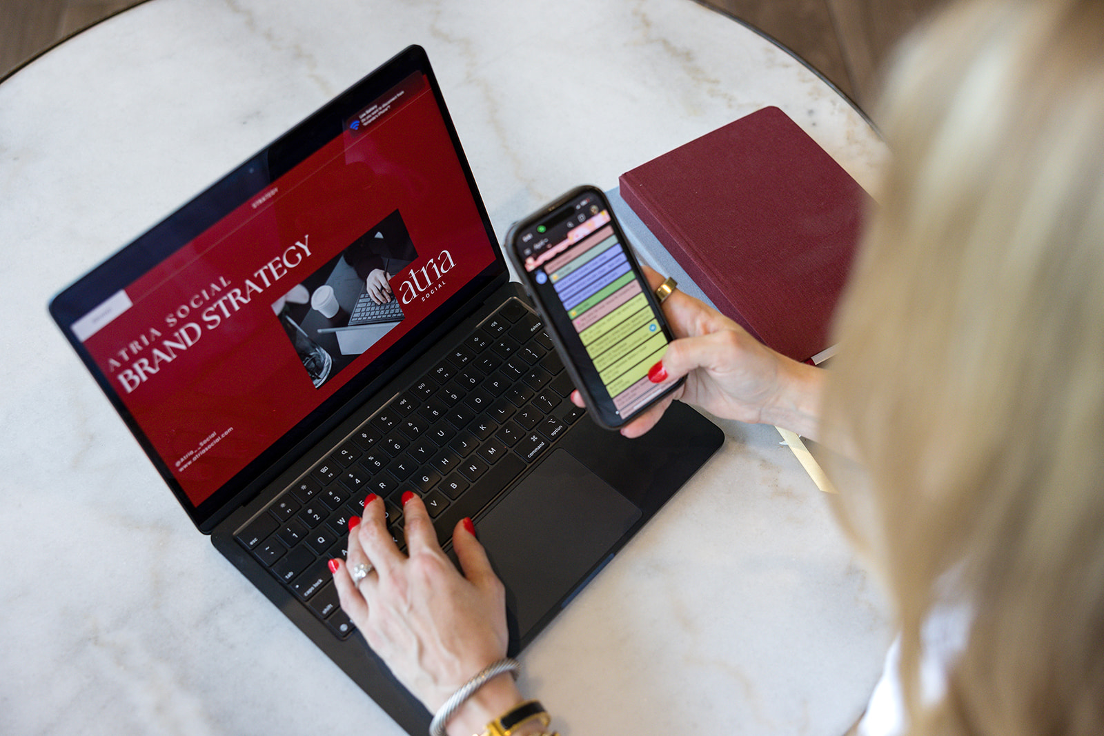 Person using a laptop and smartphone on a white surface, with a closed red notebook nearby.