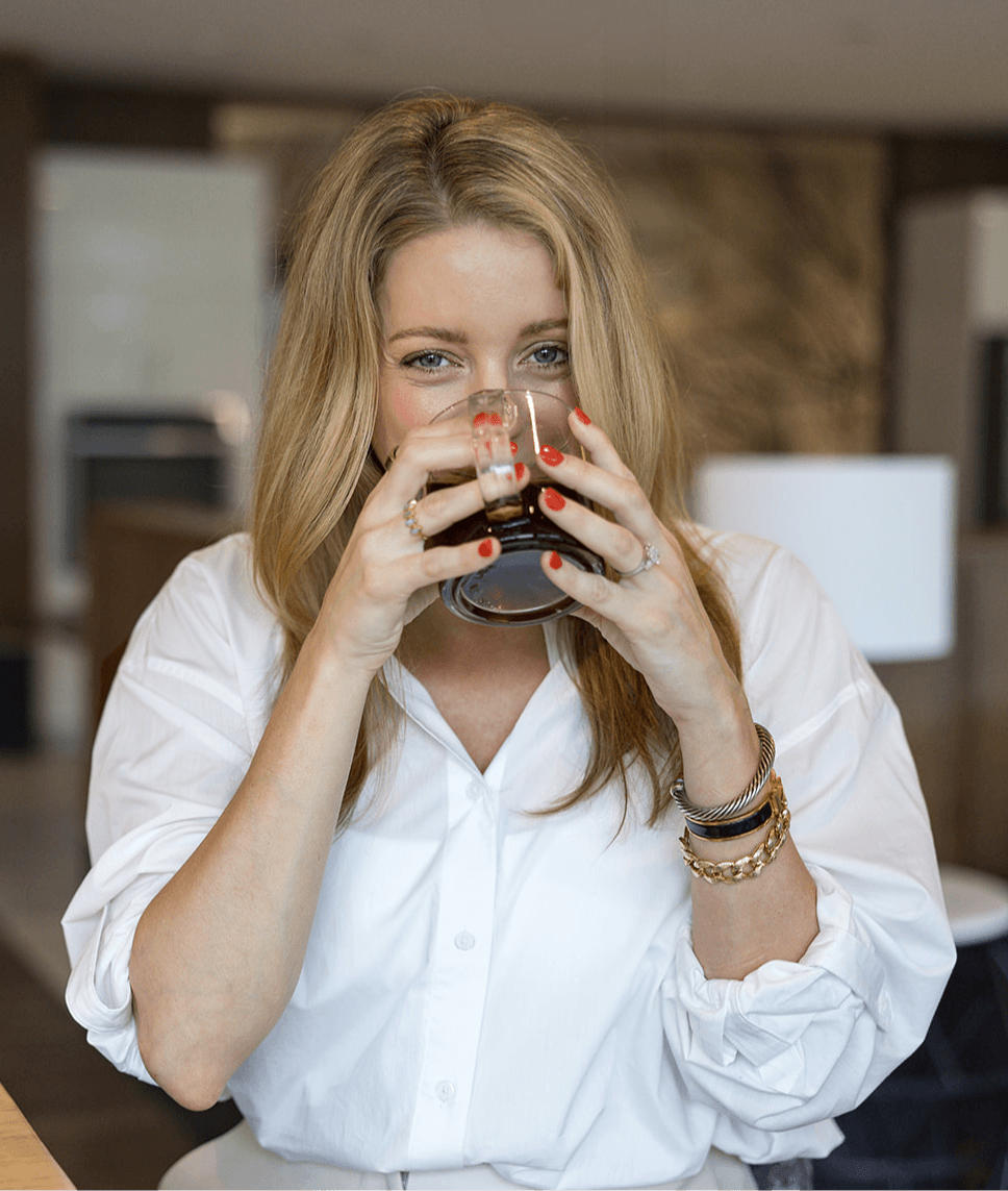 Woman with blonde hair drinking from a glass, wearing a white shirt and bracelets, in a modern indoor setting.