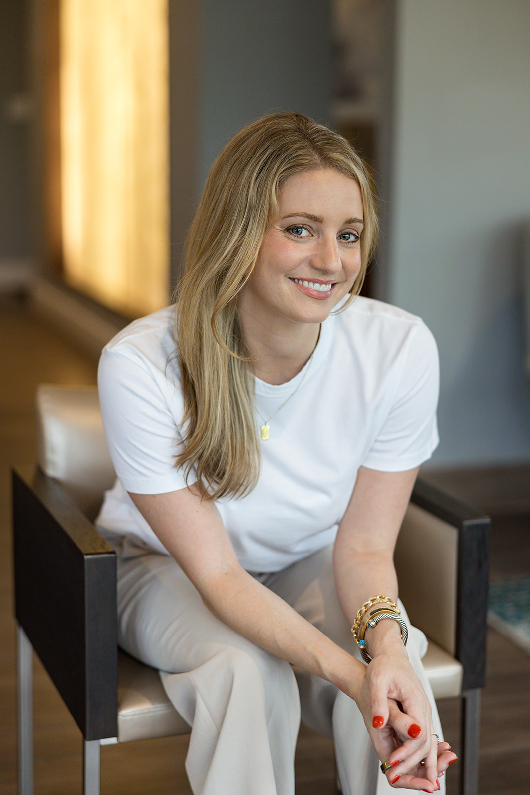 Young woman with long blonde hair smiling, sitting on a chair indoors, wearing a white t-shirt and bracelets.