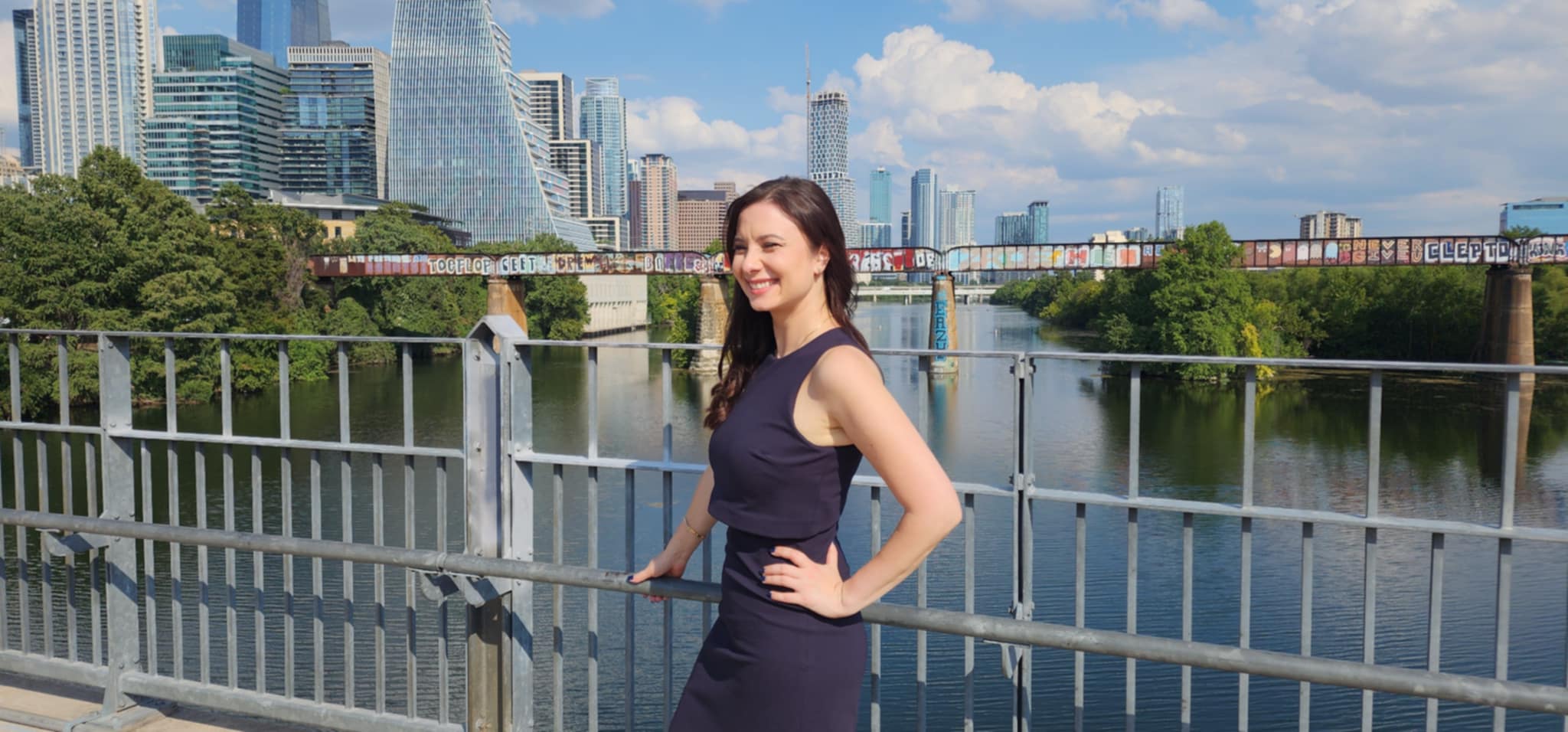 Woman in a dark dress standing by a railing near a river with city buildings in background.