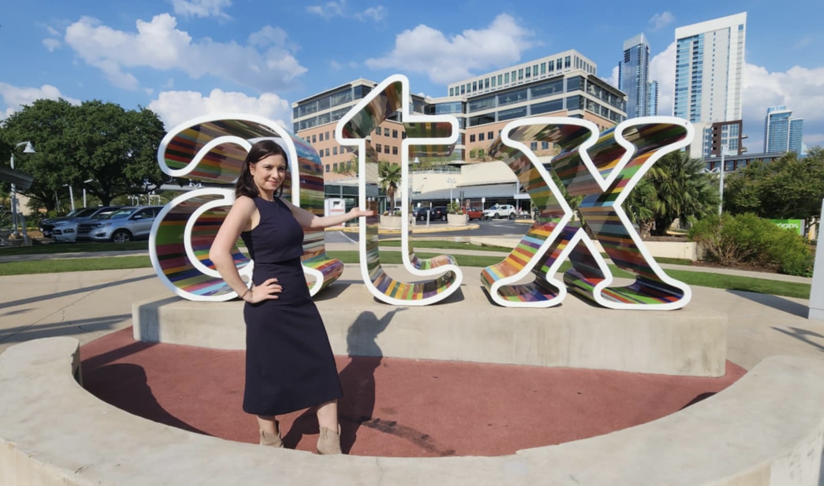 Woman in black dress standing next to large 'STL' sign outdoors with city buildings and trees in background.