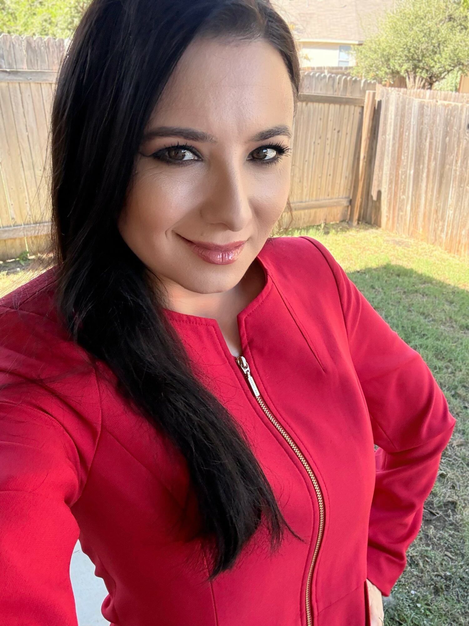 Woman with long dark hair wearing a red jacket outdoors in a backyard with a wooden fence.