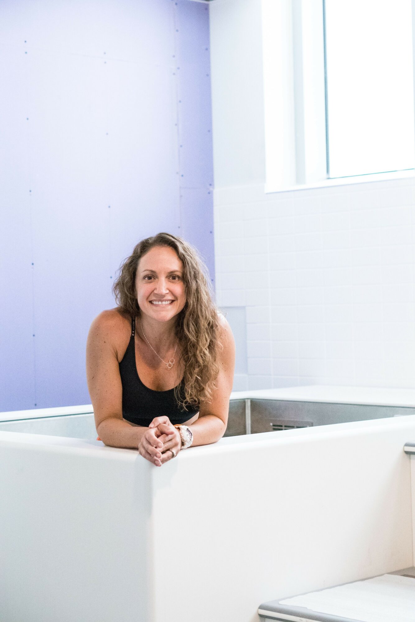 Woman with long curly hair smiling, leaning on a white railing indoors near a window.