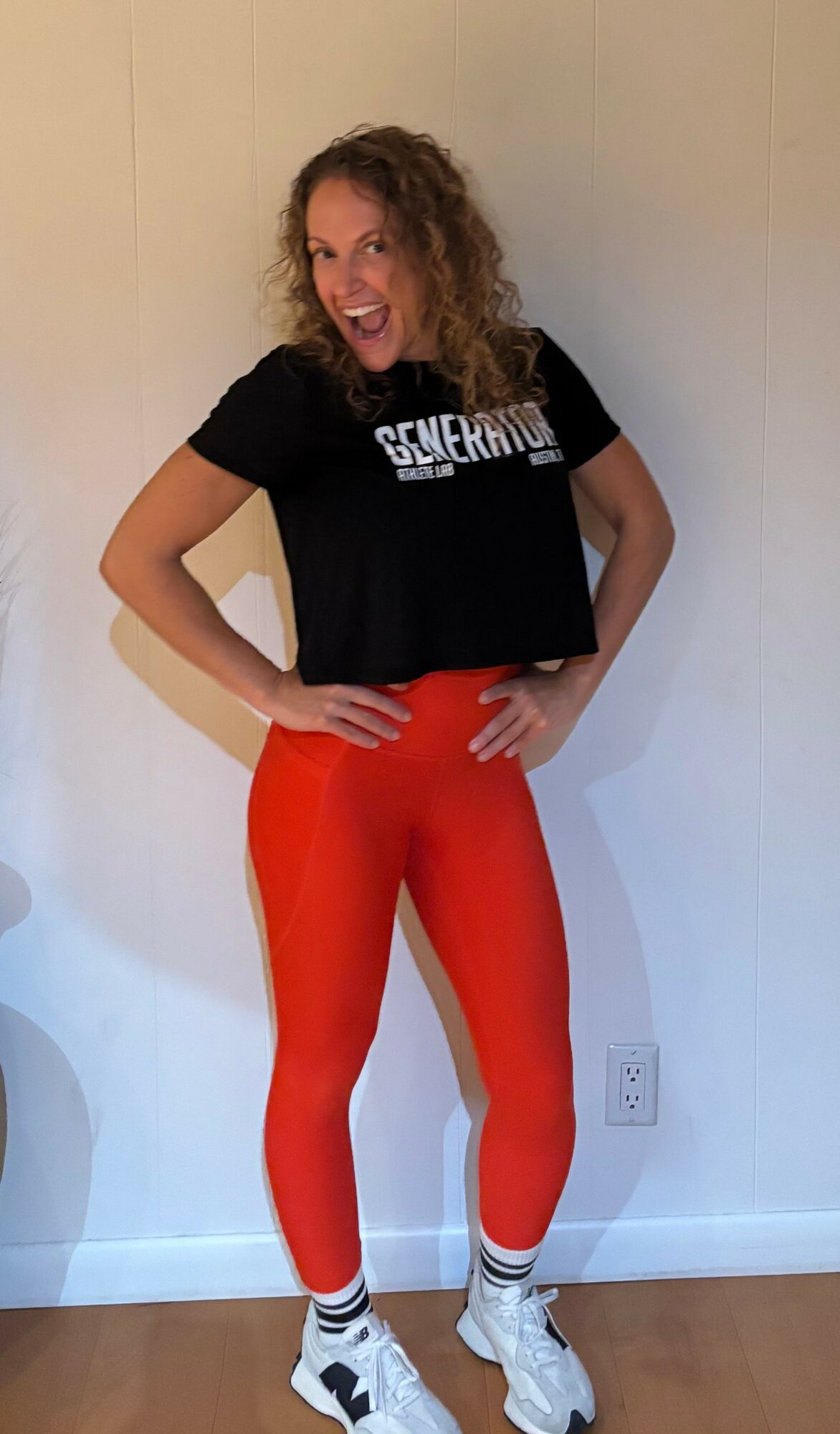 Woman with curly hair in black t-shirt and red leggings standing against a plain wall.