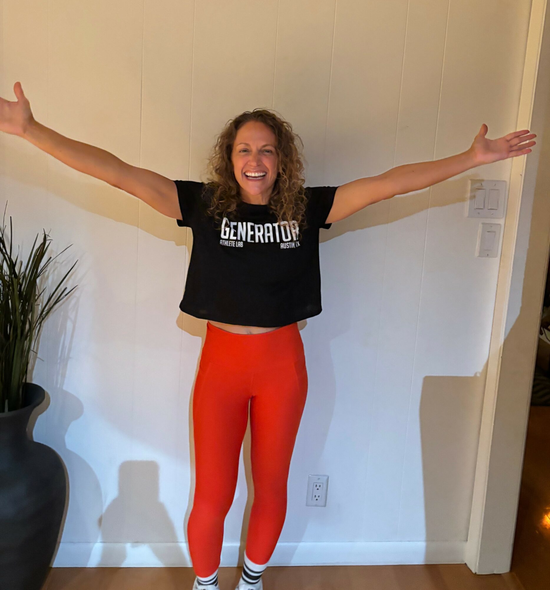 Woman with curly hair smiling, arms outstretched, standing indoors against white wall, wearing black T-shirt and red leggings.