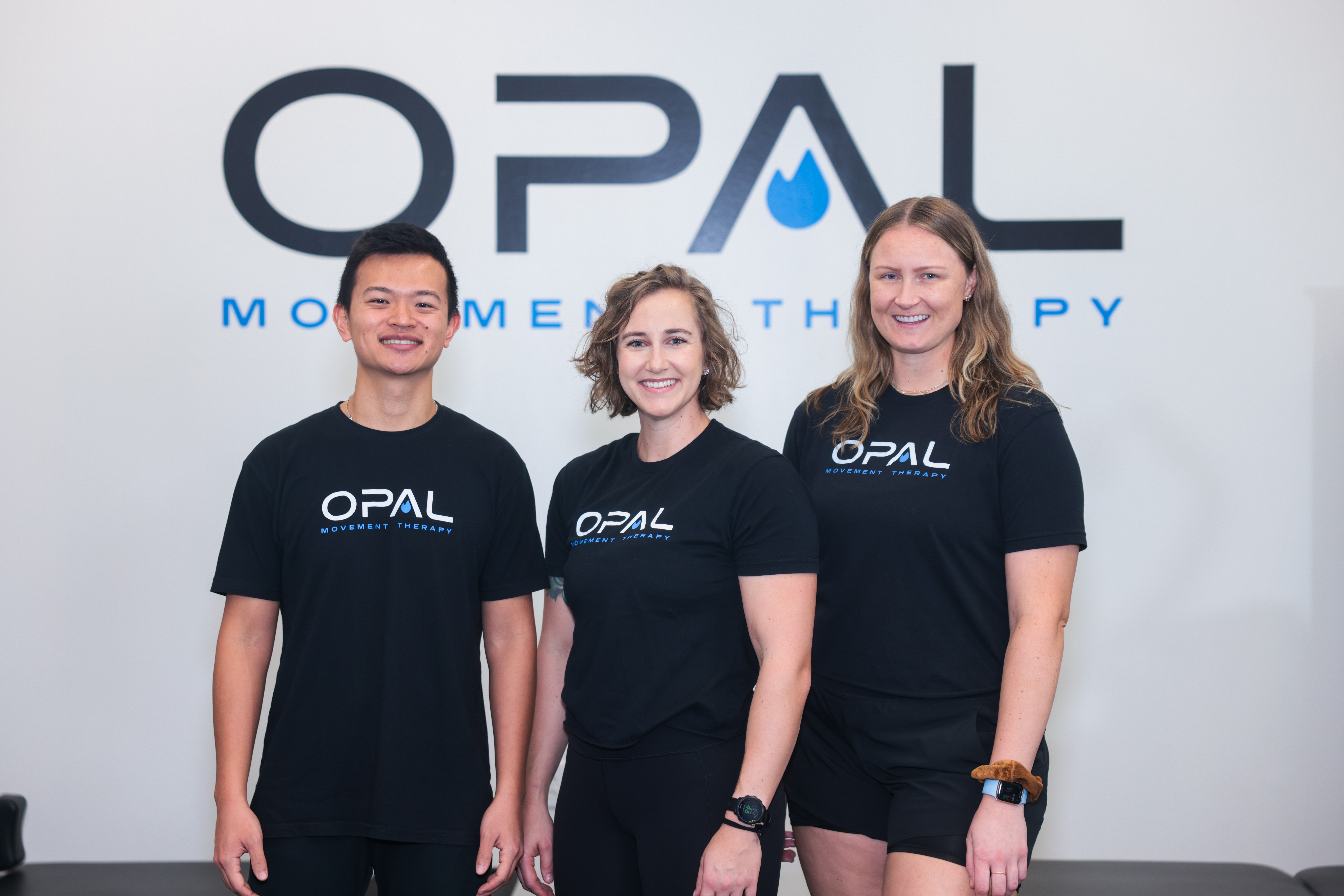 Three people wearing black OPAL shirts standing in front of a white wall with OPAL logo and text.