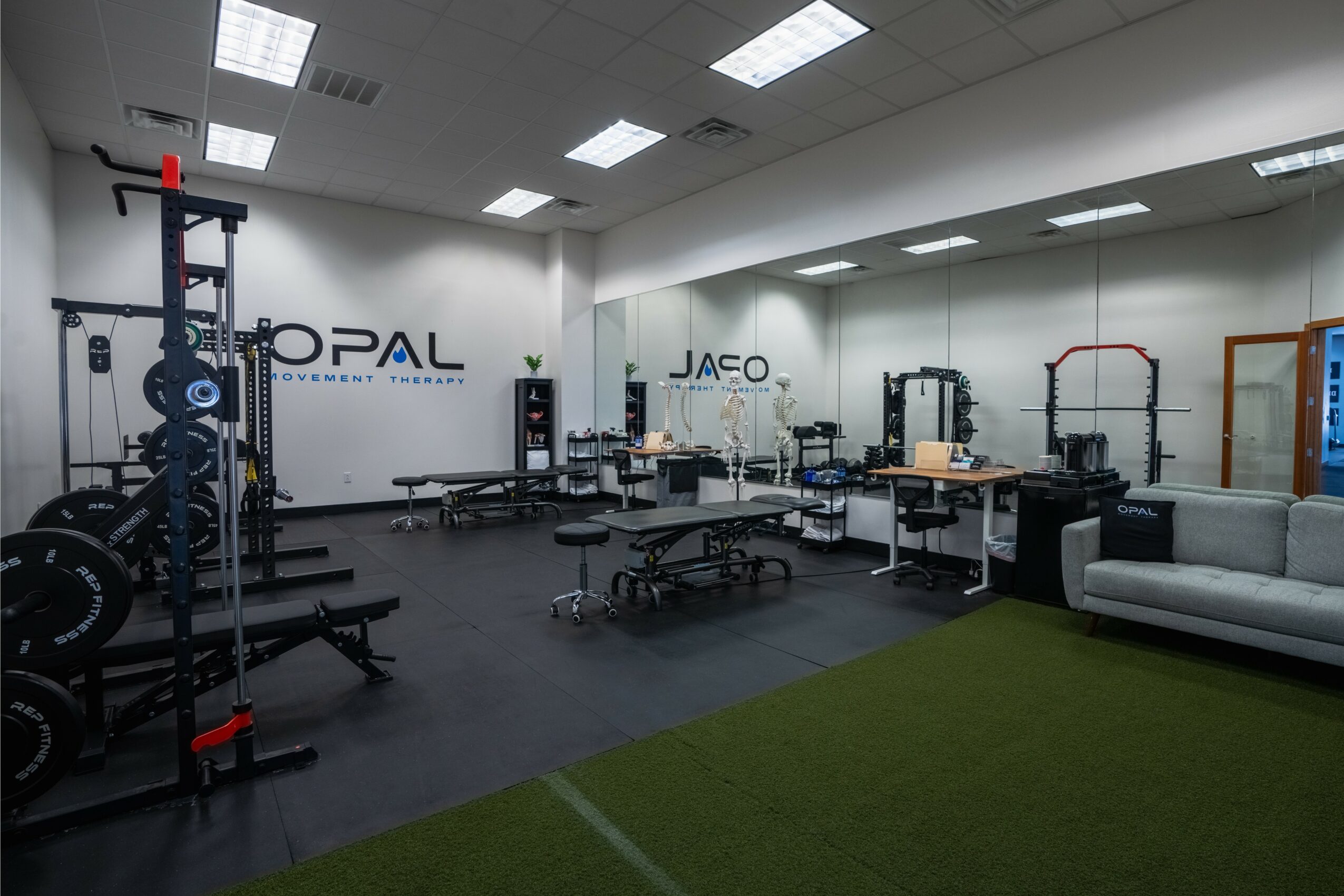 Gym with workout equipment, benches, and a mirror wall, with a green mat area and a sofa.
