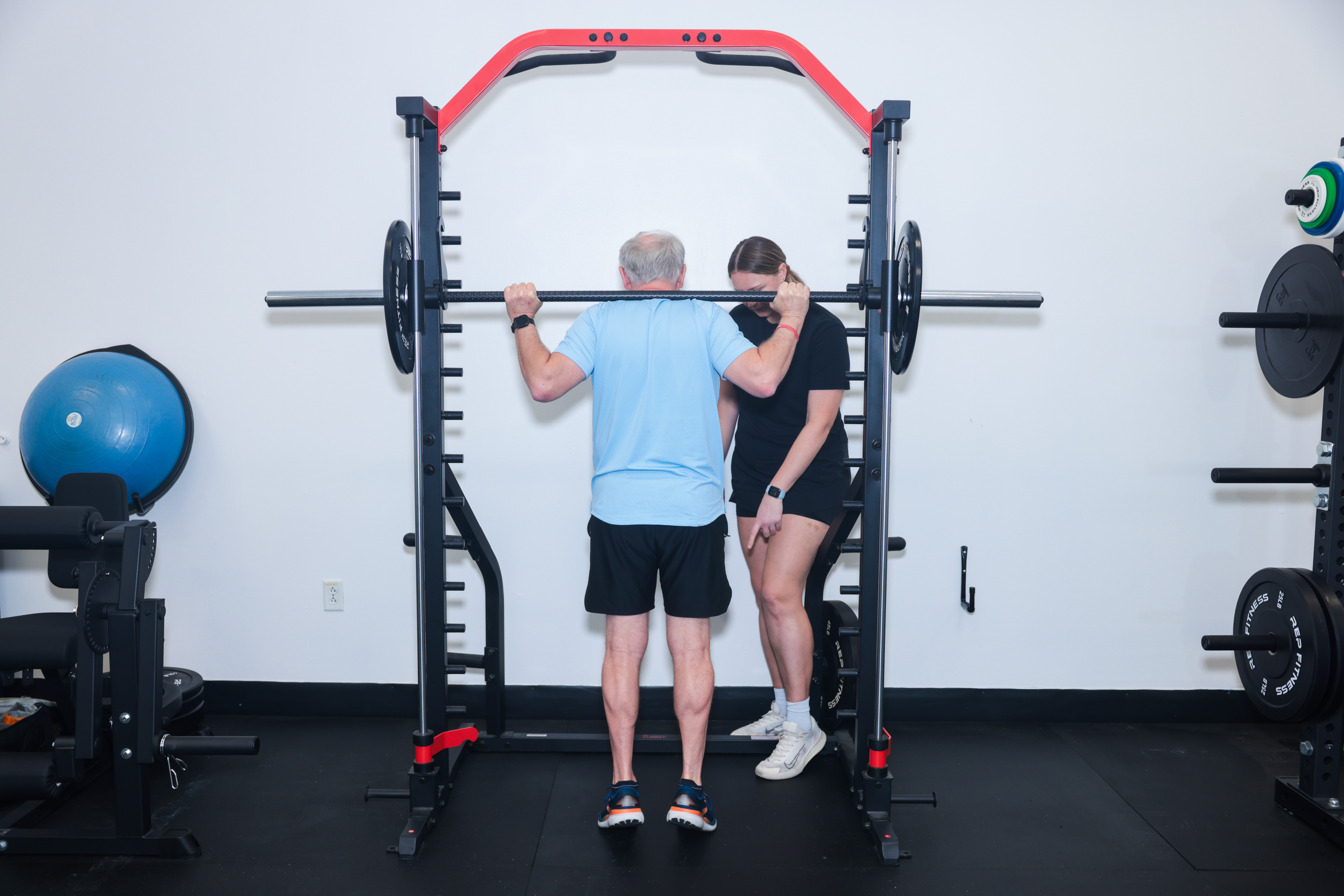 Two people in a gym, one assisting the other with a barbell on a squat rack.