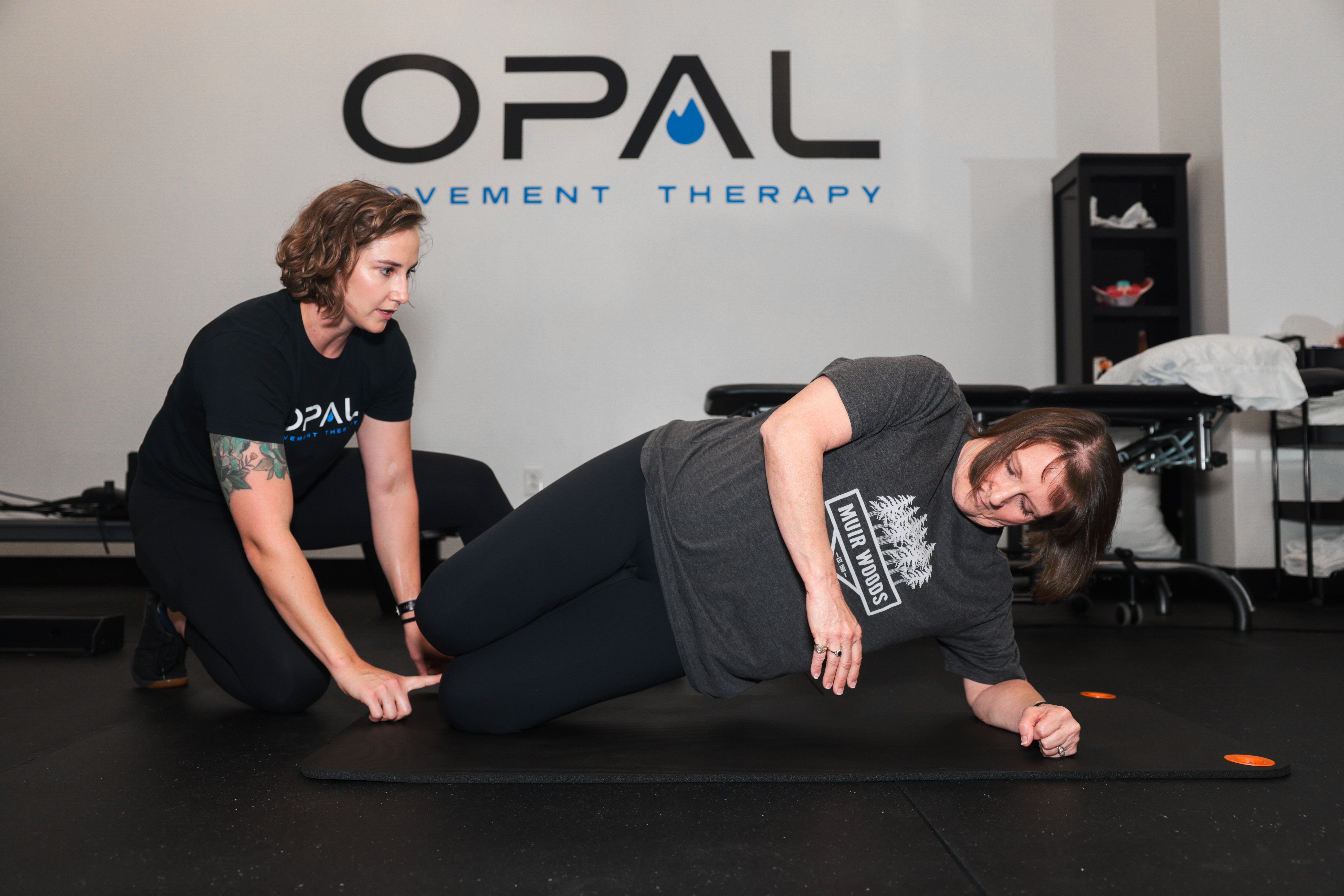 Two women in a therapy room, one assisting the other who is on her side on the floor, with a sign reading 'OPAL Movement Therapy' on the wall.