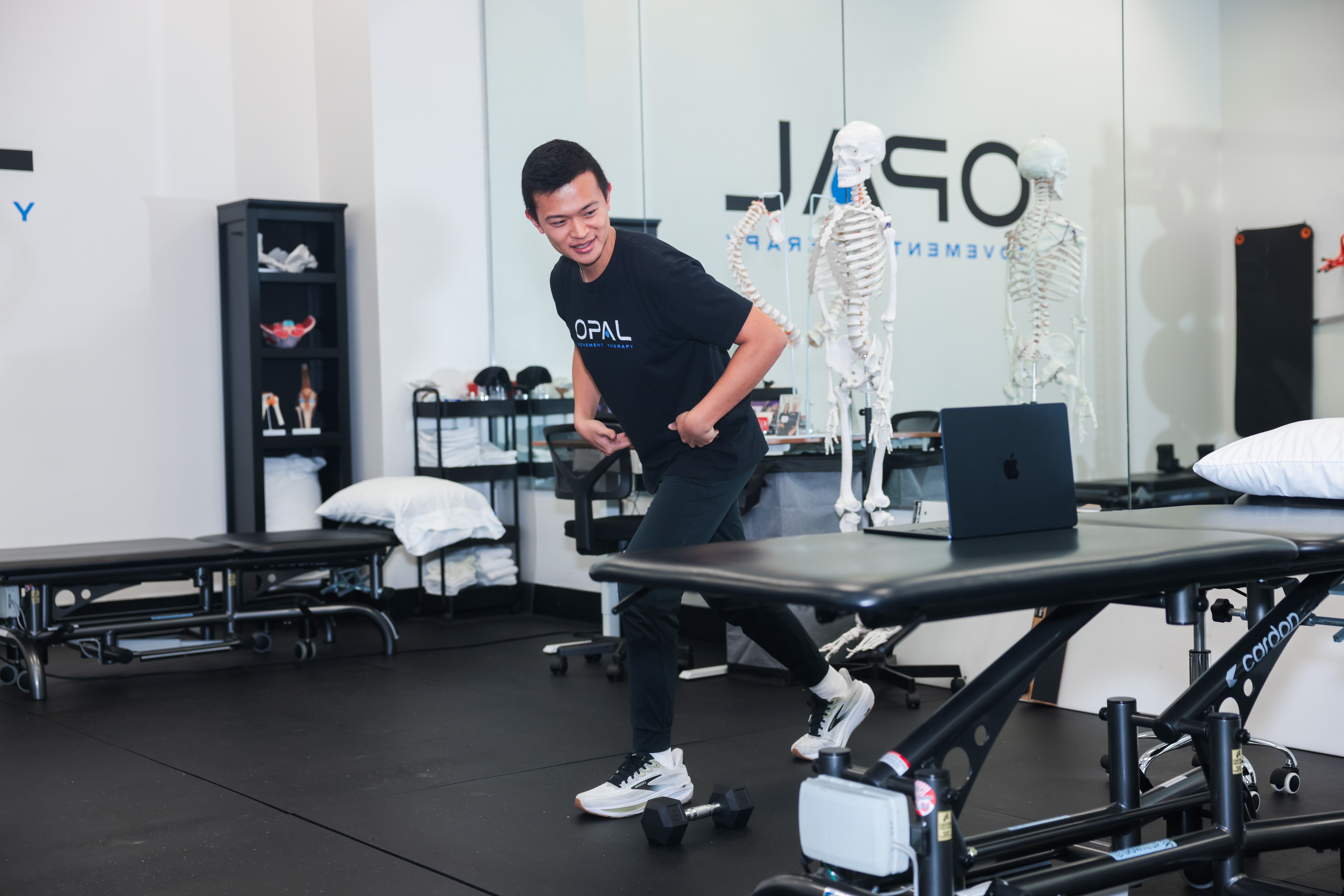 Young man in a black shirt and jeans stands in a medical training room with skeleton models and equipment.