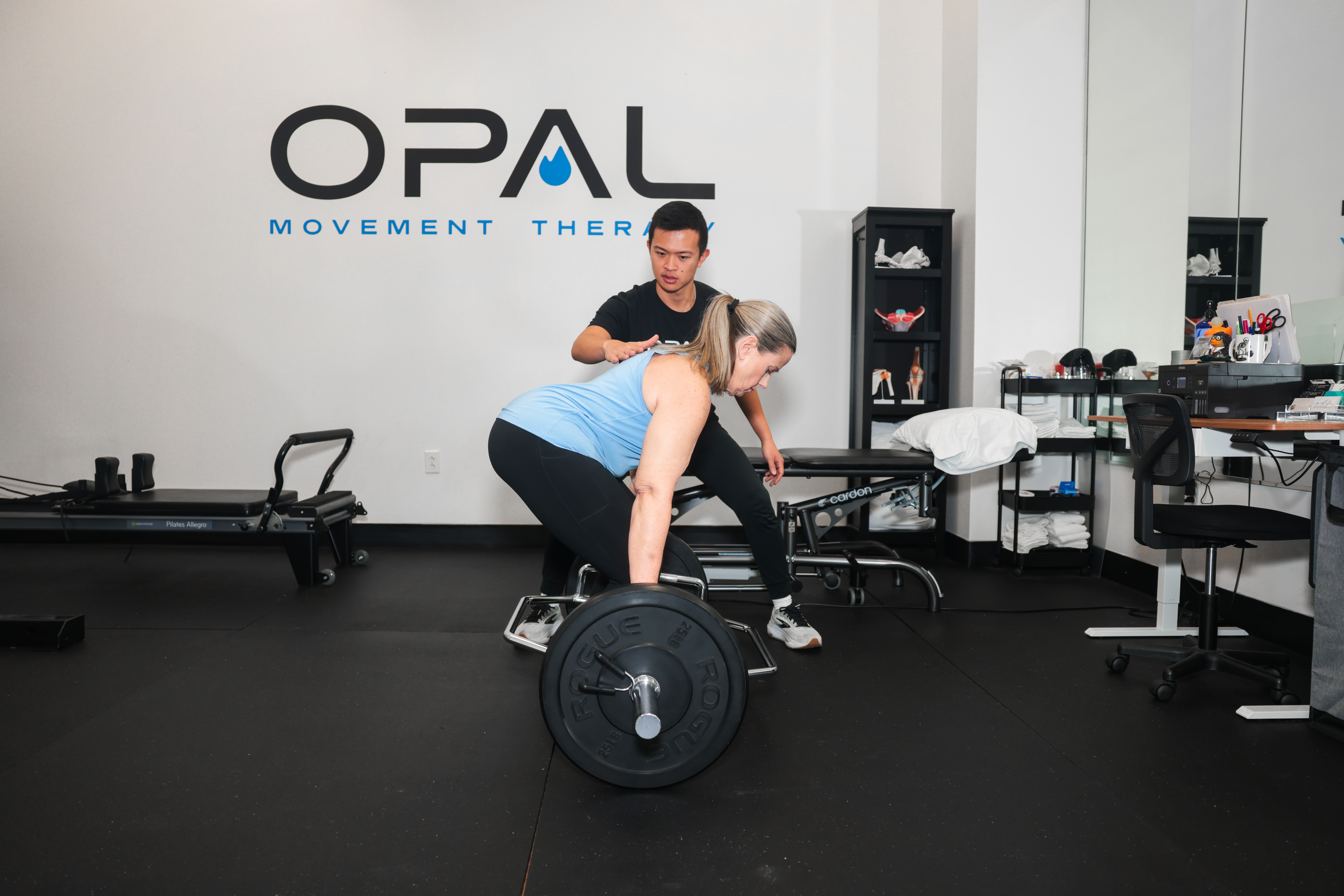Trainer assisting a woman with a deadlift exercise in a gym with OPAL logo on the wall.