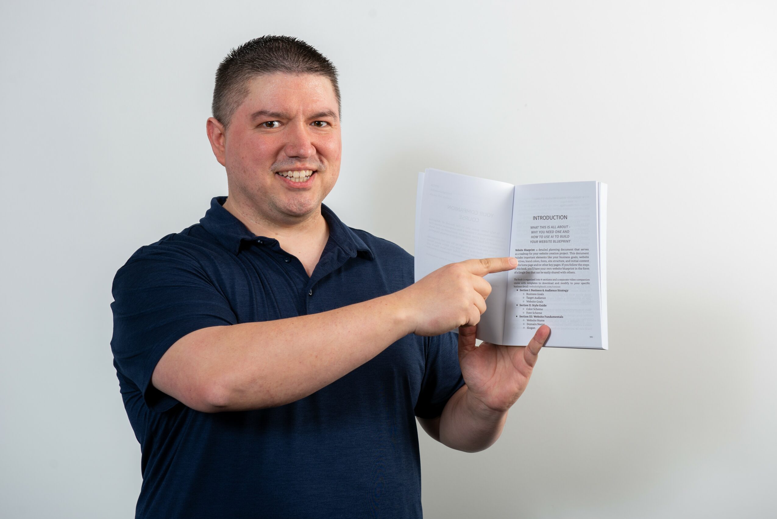 Man with short dark hair smiling, holding open book and pointing at pages, wearing a dark blue polo shirt, standing against a plain white background.