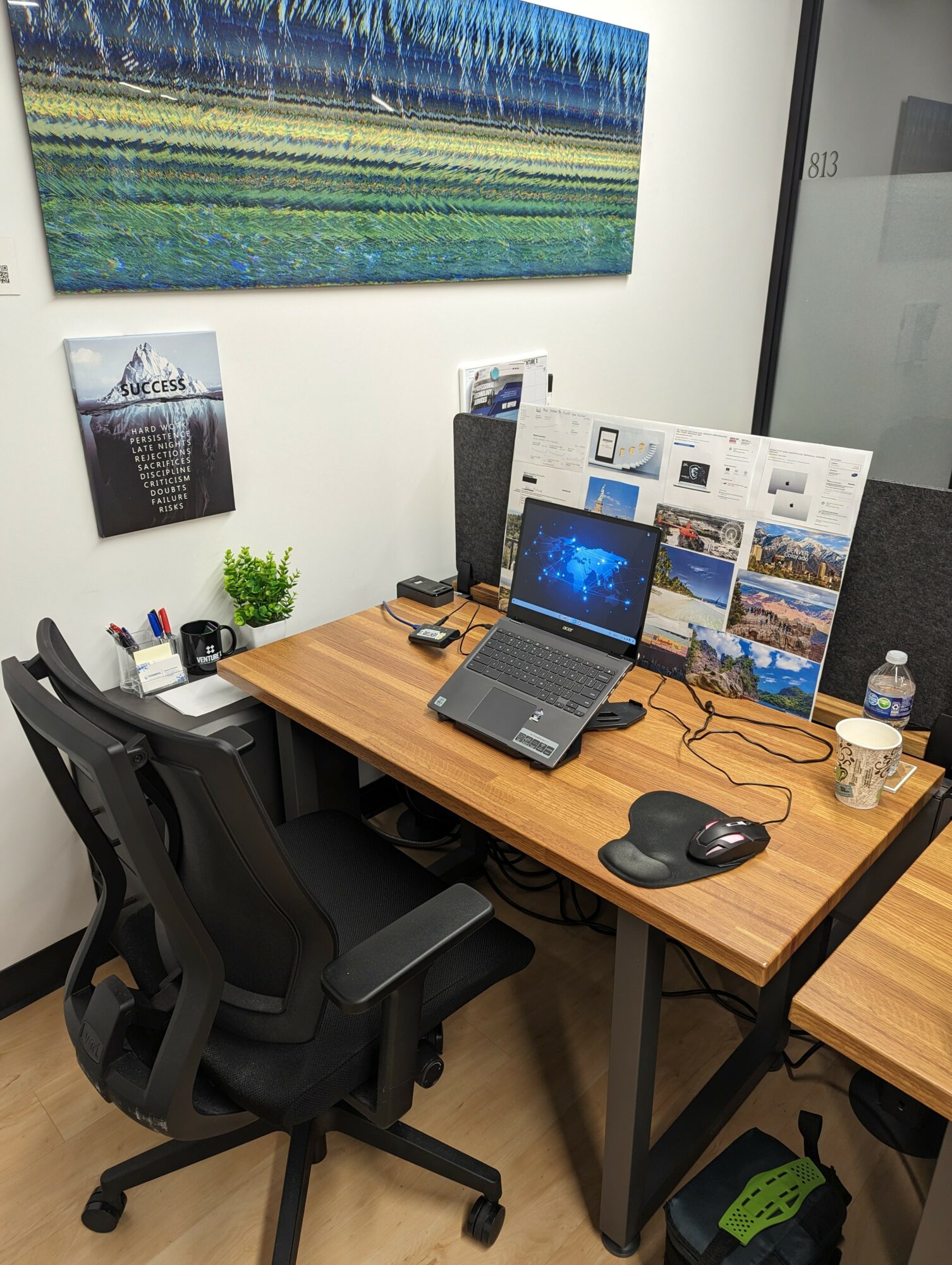 Office desk with laptop, mouse, water bottle, and brochures, surrounded by chairs and wall art.