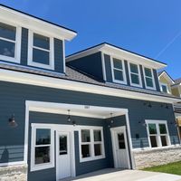 Two-story house with blue exterior, white trim, multiple windows, and a front door under a porch, against a clear blue sky.