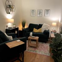 Living room with black sofa, side tables, lamps, wall art, and plants, illuminated warmly, with a wooden floor and patterned rug.