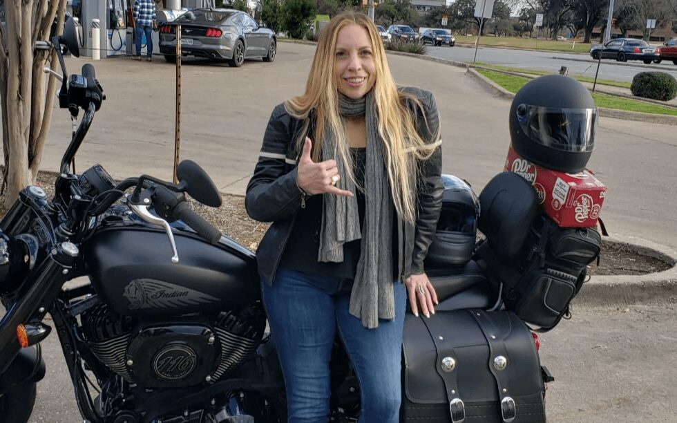 Woman with long blonde hair standing next to a black motorcycle, making a hand gesture, wearing a black leather jacket and gray scarf.