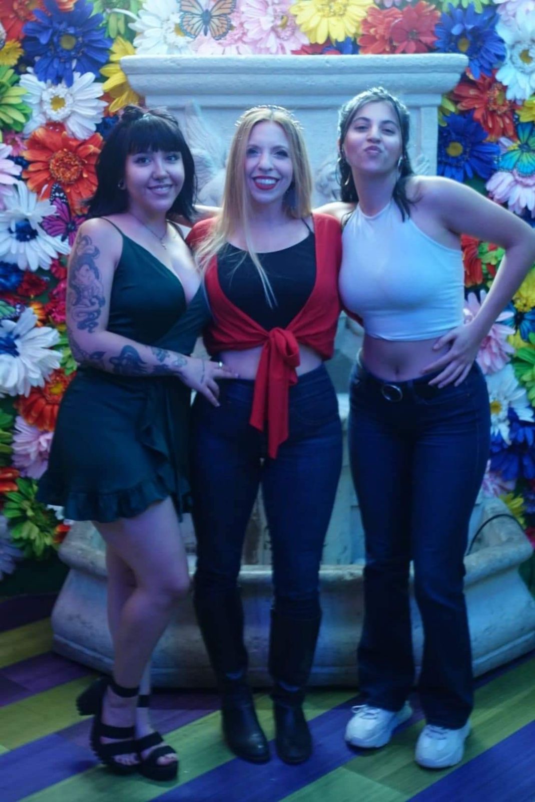 Three women standing in front of a colorful flower backdrop, smiling, with arms around each other.