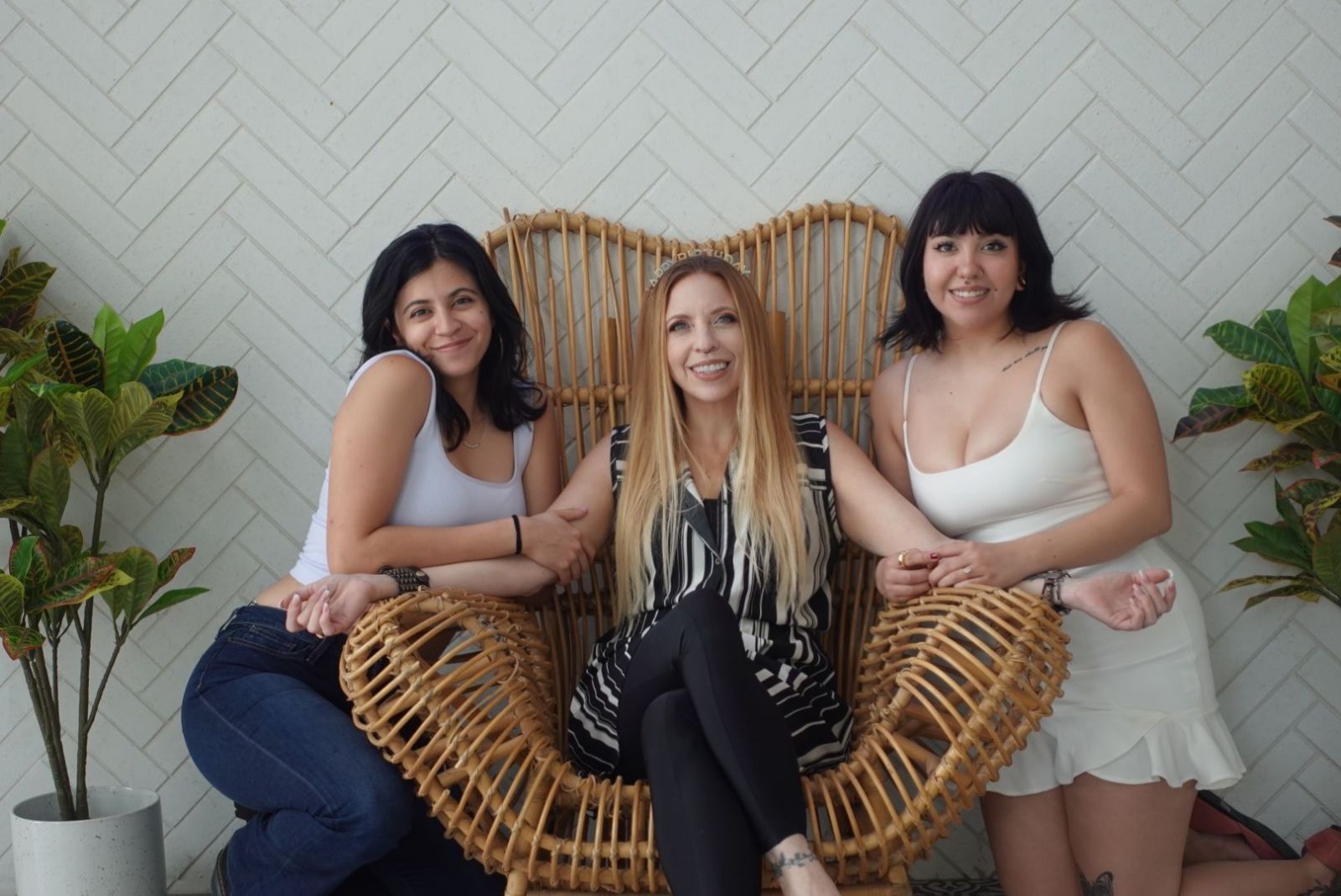 Three women sitting on a wicker heart-shaped chair, smiling, with plants on each side and a white wall background.