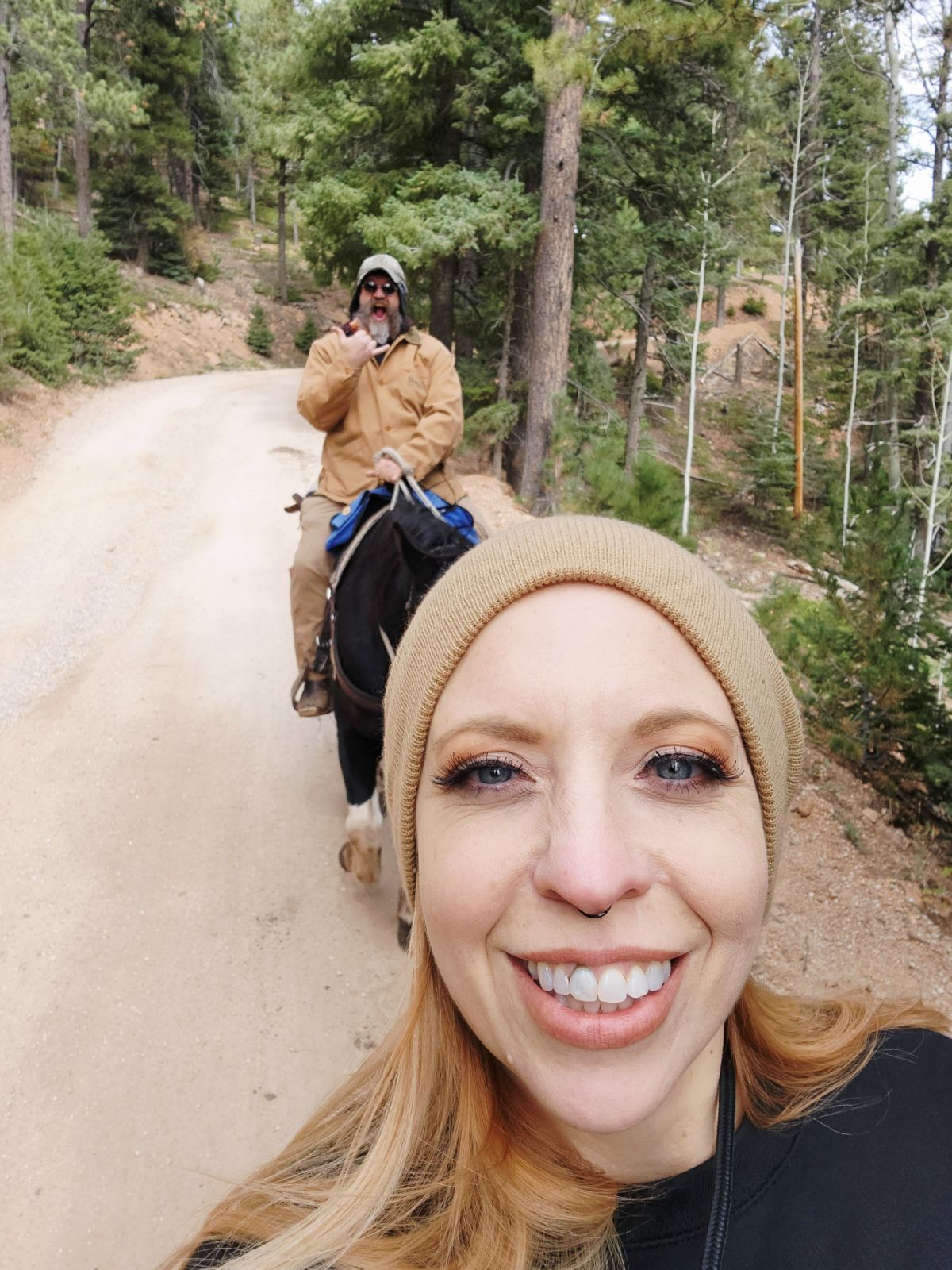 Two women on a dirt trail in a forest, one smiling in the foreground, the other taking a photo in the background.
