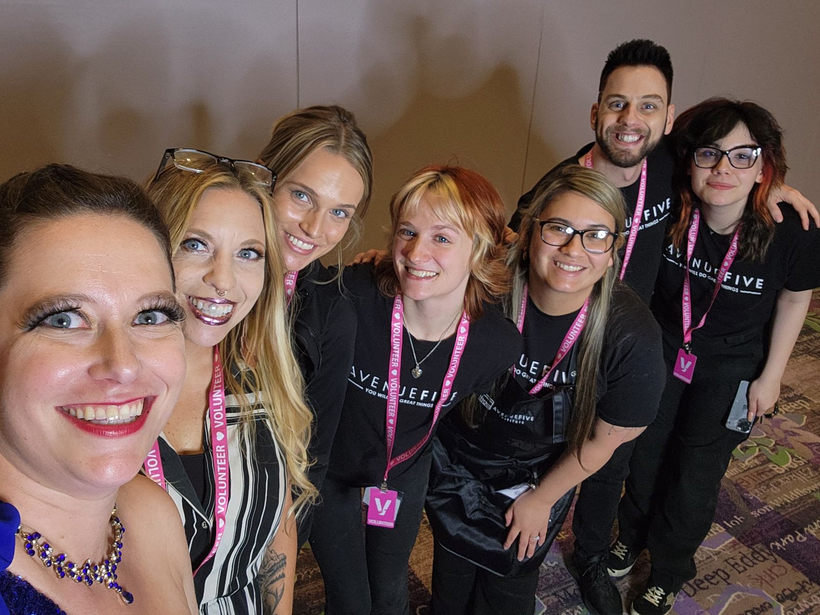 Group of seven smiling people standing together indoors, some wearing black shirts and pink lanyards, posing for a photo.