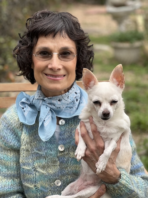Woman with short curly hair and glasses holding a small white dog outdoors, greenery in background.