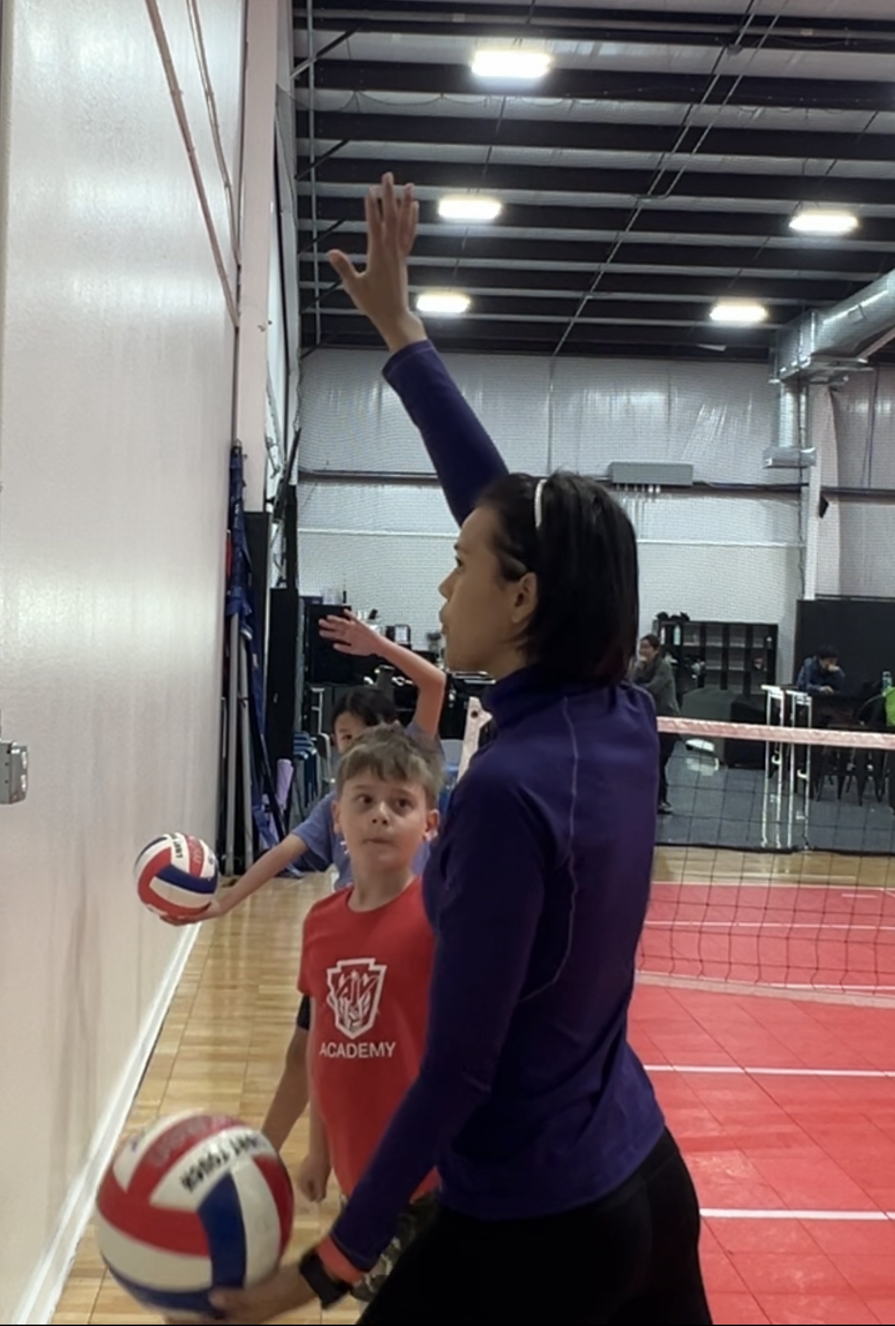 Woman with dark hair in a purple jacket raises her hand, two children with volleyballs stand nearby in a gym.