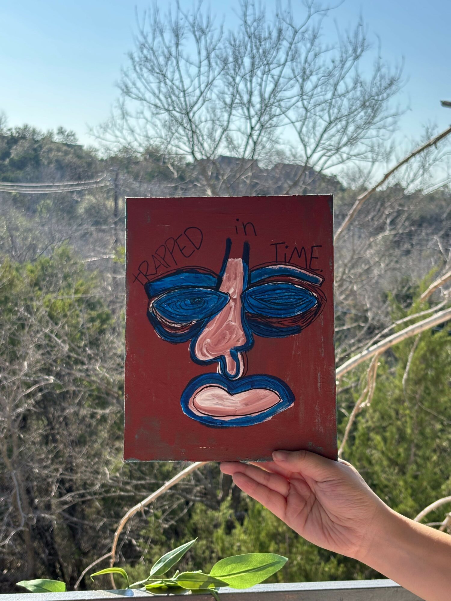 Person holding a colorful painted sign with a face, trees and sky in background.
