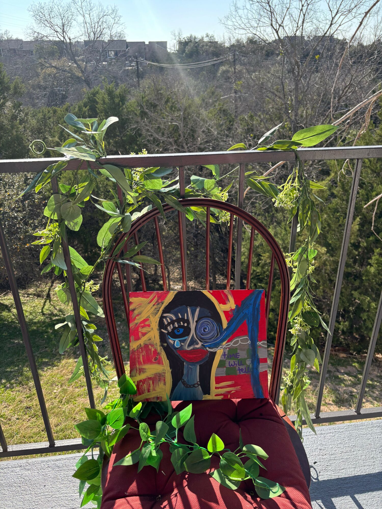 Colorful pillow with abstract face design on a metal chair outdoors, surrounded by green plants and a railing.