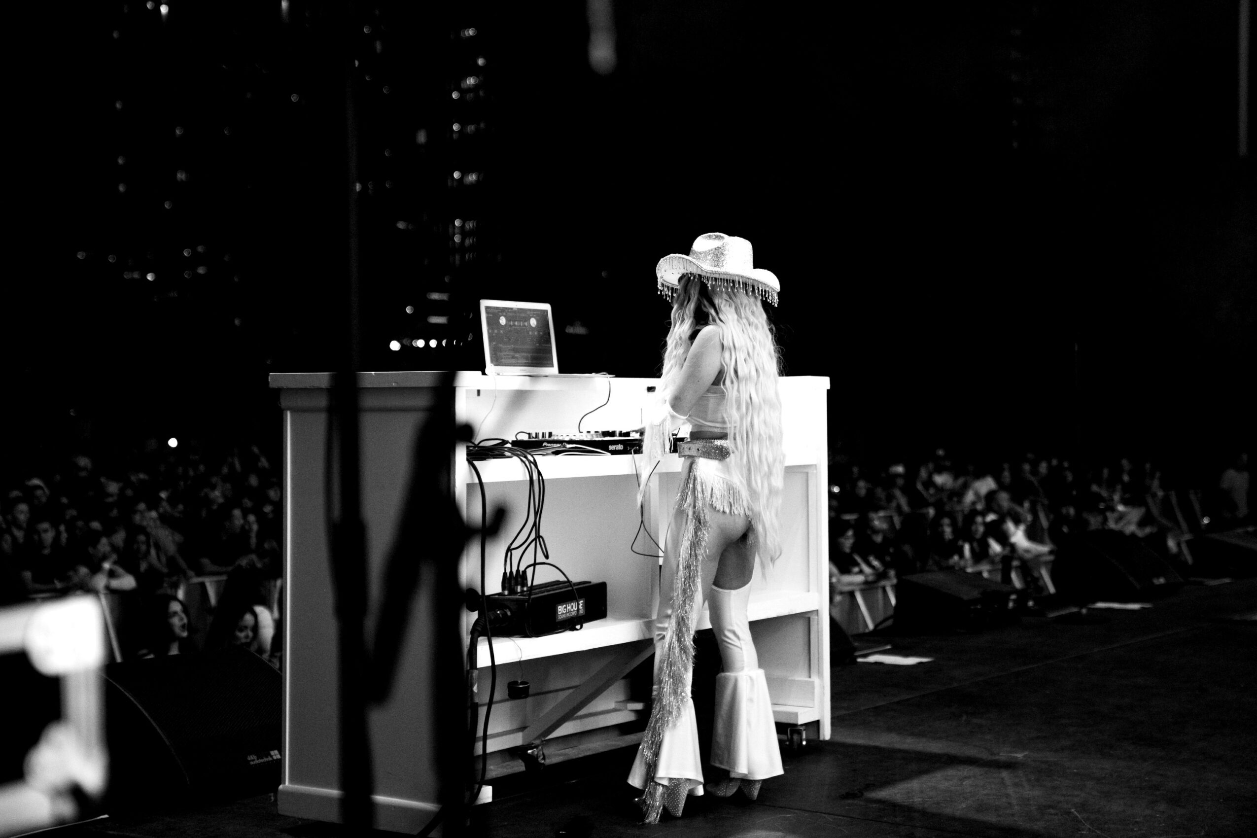 Person wearing a hat and light clothing stands behind a DJ setup on stage at night.