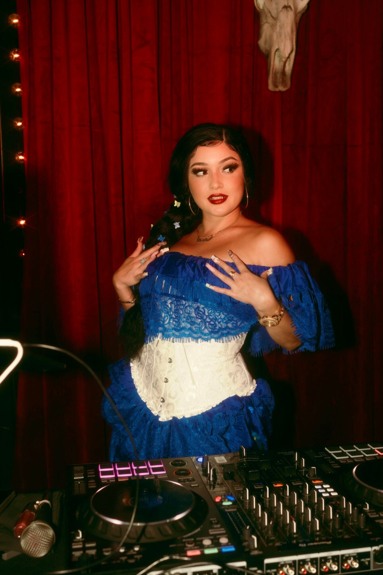 Woman with dark hair and makeup, wearing a blue and white off-shoulder dress, standing in front of red curtains.