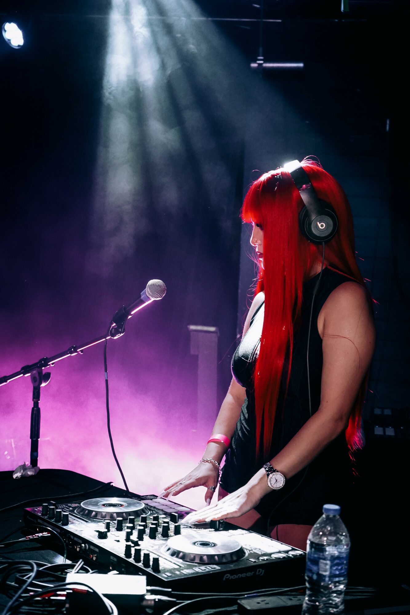 Female DJ with long red hair wearing headphones, operating DJ equipment on stage with purple and blue lighting.