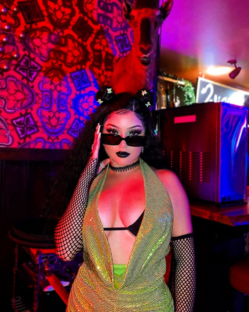 Person with dark hair, sunglasses, black lipstick, fishnet gloves, and a glittery outfit in a colorful, dimly lit setting.
