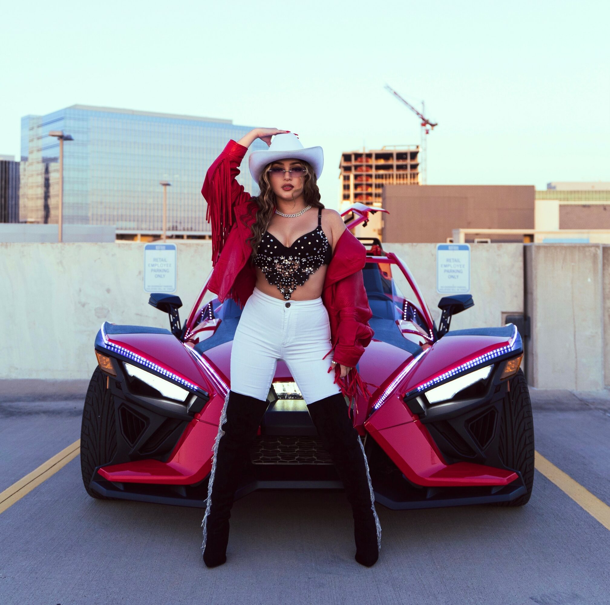 Woman in red jacket, white hat, and sunglasses standing in front of a red sports car on a rooftop parking lot.