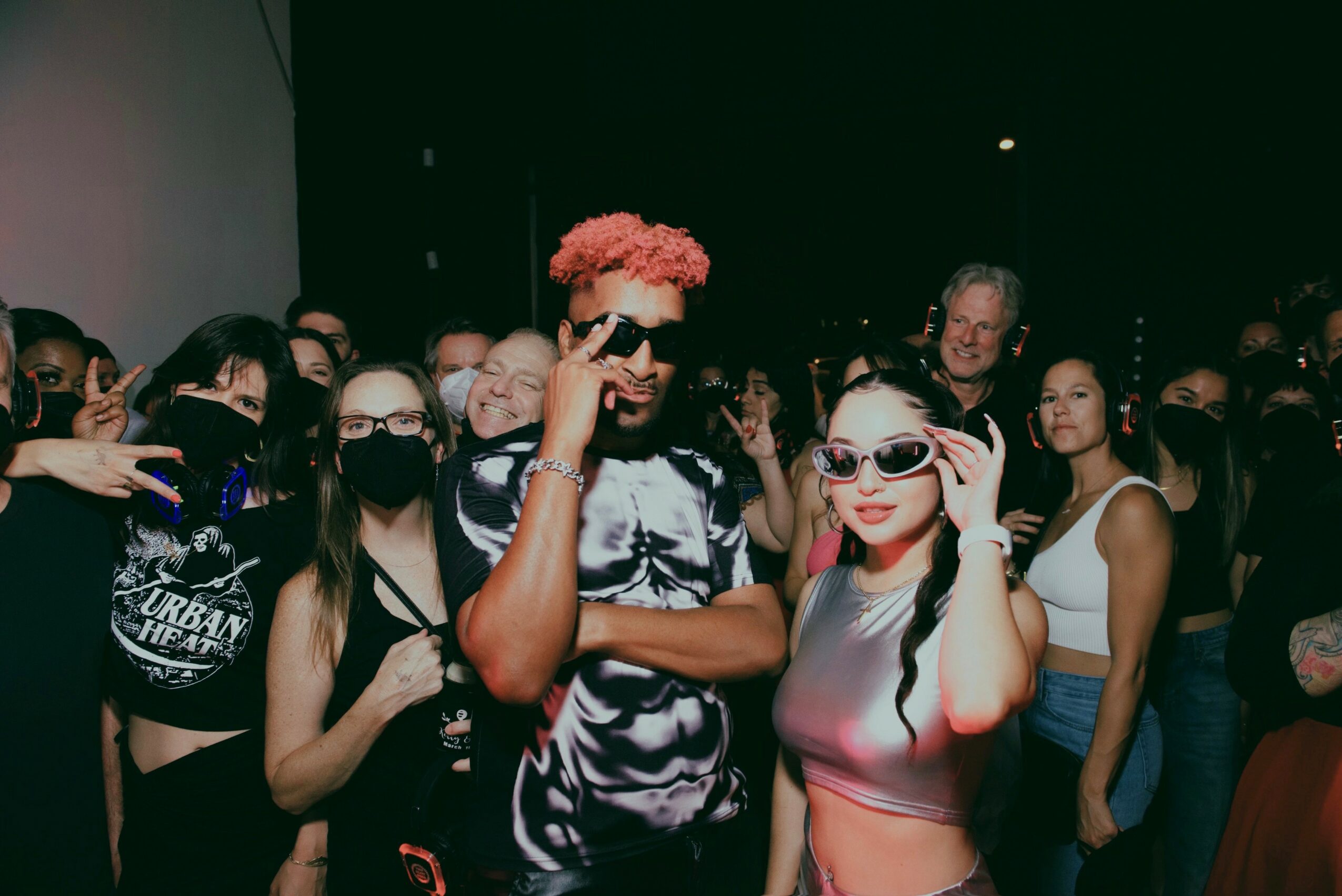 Group of people at night, some wearing sunglasses and masks, posing for a photo with a person in the center.