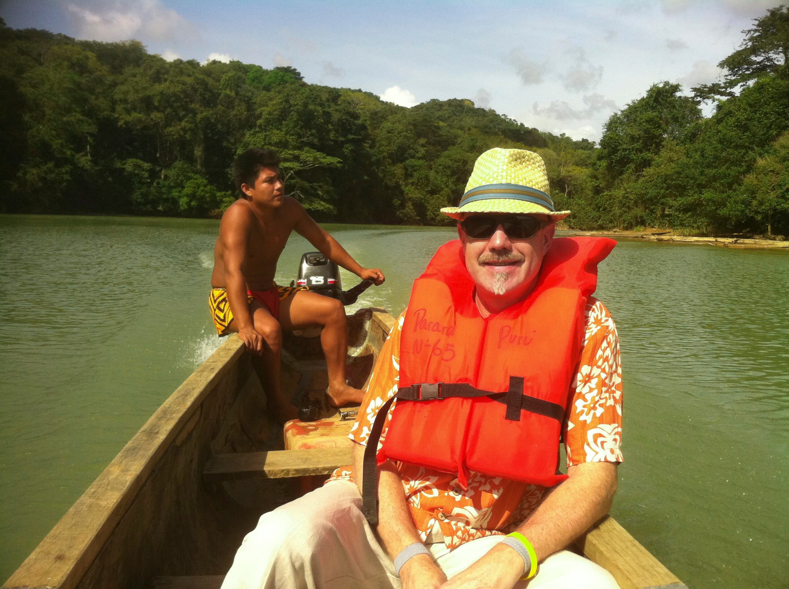 Older man wearing a hat, sunglasses, and life jacket sitting in a boat with a young boy steering on a lake surrounded by trees.