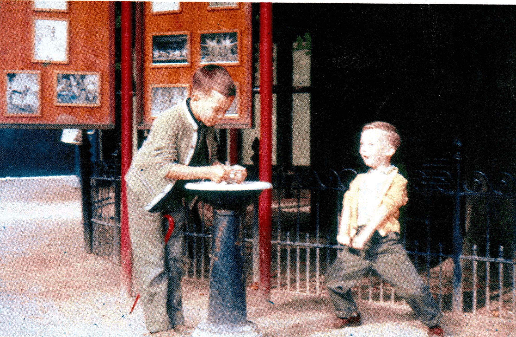 Two children, one leaning on a stand and the other standing, outdoors near a fence and display boards.