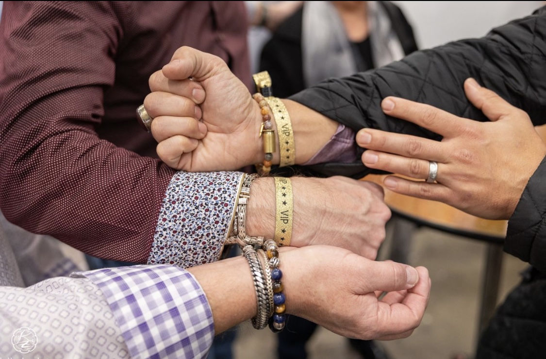 Multiple hands and arms engaged in a handshake or physical interaction, wearing various bracelets and rings.