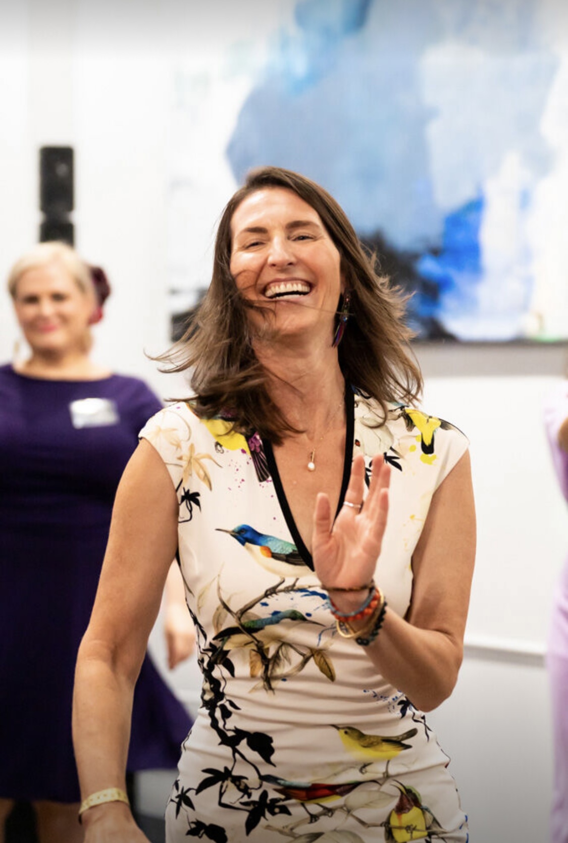 Woman smiling and waving, wearing a sleeveless dress with bird patterns, in an indoor setting with a blurred background.