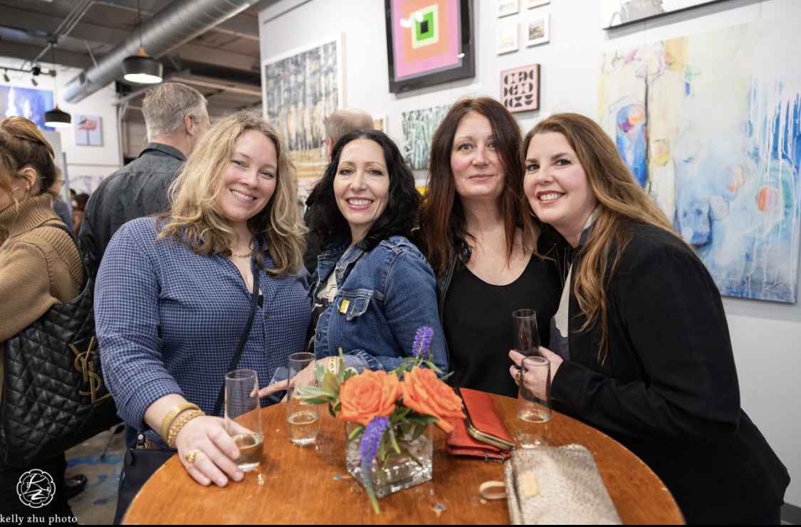 Four women smiling and holding drinks around a table with flowers at an art gallery.